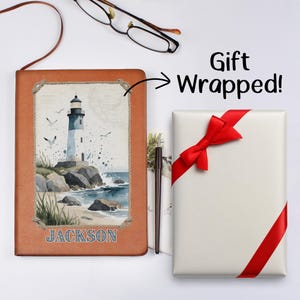 Op de afbeelding: Een bruin leren notitieboek met een vuurtorenillustratie en de naam "JACKSON" staat naast een cadeauverpakking met een rode strik. De tekst "Gift Wrapped!" is ook zichtbaar.