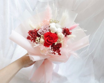 May include: A bouquet of preserved flowers in a pink paper wrapping. The bouquet includes a red rose, a pink rose, white cotton, and red dried flowers.