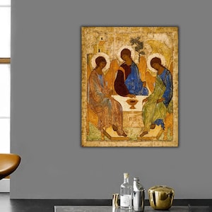 May include: A rectangular wall art print featuring a reproduction of the Rublev Trinity icon. The artwork depicts three figures with halos seated around a table with a chalice, rendered in muted gold, blue, green, and brown tones on a textured background.