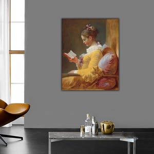 Puede incluir: Un óleo enmarcado que representa a una persona con un vestido amarillo y cuello de volantes blancos, sentada y leyendo un libro abierto. La persona lleva el cabello recogido con un lazo y se apoya en cojines suaves y texturizados en una habitación cálida.