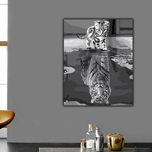 May include: Black and white artwork of a kitten standing in water, with its reflection appearing as a tiger. The painting is on a canvas and is displayed on a gray wall. The artwork is in a room with a window and a chair.