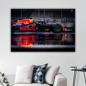 May include: A red and black race car with the words "Red Bull" on the side, reflecting in a puddle of water. The car is parked in a garage with a metal grid wall behind it.