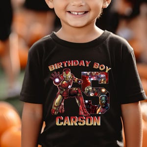 May include: A black t-shirt with the text "BIRTHDAY BOY" and the name "CARSON" printed on it. The shirt features a graphic of Iron Man and the number 5. The boy is smiling.