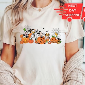 May include: A cream-colored short-sleeve t-shirt featuring a graphic print of Disney characters dressed in skeleton costumes sitting among orange pumpkins and black spiderwebs. The design includes Goofy, Donald, Mickey, and Minnie Mouse.