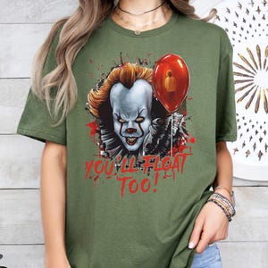 May include: Green t-shirt with a graphic of Pennywise the clown from the movie "IT" with a red balloon. The text "You'll Float Too!" is printed below the graphic.