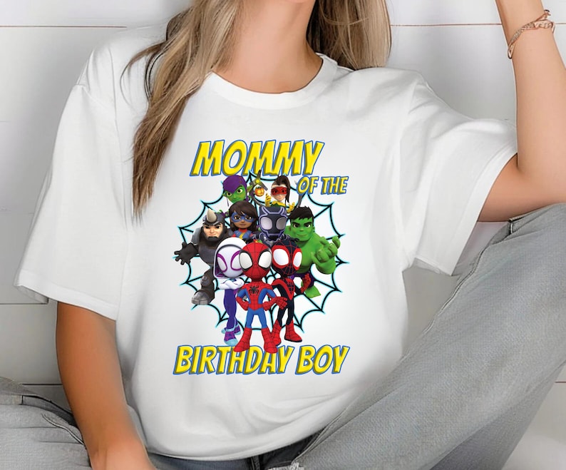 Puede incluir: Camiseta blanca con un gr&aacute;fico colorido de superh&eacute;roes de dibujos animados, incluyendo a Spider-Man y Hulk. El texto "MOMMY OF THE BIRTHDAY BOY" est&aacute; impreso en amarillo con un contorno azul. Un dise&ntilde;o alegre y festivo.