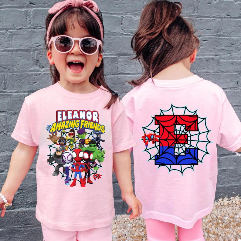 May include: A light pink short-sleeved t-shirt featuring a graphic print of various superhero characters and the text "Eleanor and Amazing Friends" on the front, with a large letter "E" in a spiderweb design on the back of the garment.
