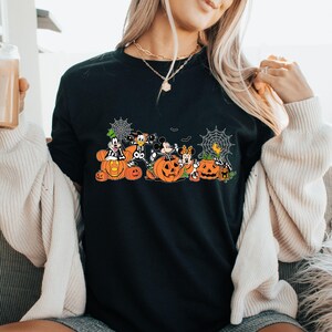 May include: A black t-shirt featuring a graphic print of cartoon characters dressed in skeleton costumes, sitting inside and around orange jack-o'-lantern pumpkins, accented with white spiderwebs and small bats against a dark background.