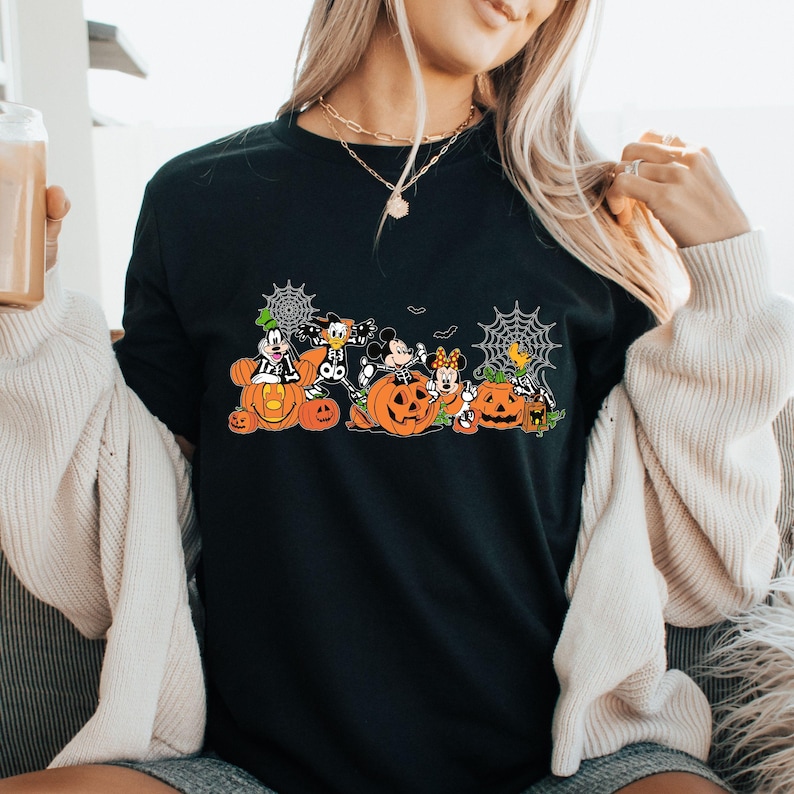 May include: A black t-shirt featuring a graphic print of cartoon characters dressed in skeleton costumes, sitting inside and around orange jack-o'-lantern pumpkins, accented with white spiderwebs and small bats against a dark background.