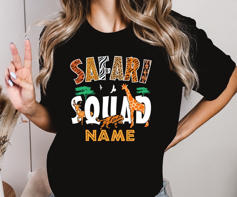 以下が含まれることがあります： 「サファリ・スクワッド」というカラフルなグラフィックがプリントされた黒いTシャツ。キリン、トラ、鹿が描かれています。「NAME」という文字がグラフィックの下にあります。