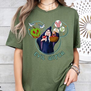May include: Green t-shirt featuring a graphic of the Evil Queen from Snow White. The graphic depicts the Evil Queen holding a basket of apples and a Mickey Mouse head silhouette with birds and apples. The text "Evil Witch" is printed below the graphic.