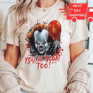 May include: A white t-shirt with a graphic of Pennywise the clown from the movie "IT". The graphic shows Pennywise with a red balloon and the text "You'll Float Too!"