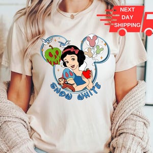 May include: A white t-shirt featuring a graphic of Snow White, a bird, and an apple. The graphic is in a blue and white Mickey Mouse ear shape. The text "Snow White" is printed below the graphic.