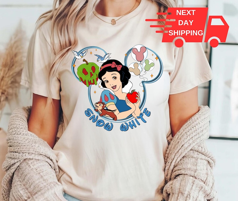 May include: A white t-shirt featuring a graphic of Snow White, a bird, and an apple. The graphic is in a blue and white Mickey Mouse ear shape. The text "Snow White" is printed below the graphic.