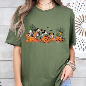 May include: An olive green t-shirt featuring a graphic print of cartoon characters dressed in skeleton costumes, sitting inside and around orange jack-o'-lantern pumpkins, accented with black spider web illustrations and small bats.