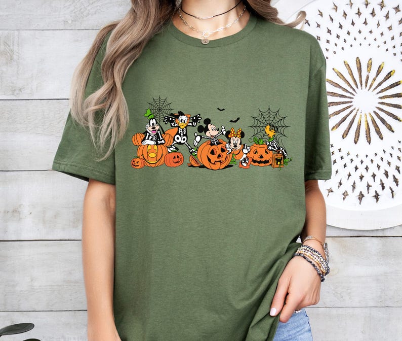 May include: An olive green t-shirt featuring a graphic print of cartoon characters dressed in skeleton costumes, sitting inside and around orange jack-o'-lantern pumpkins, accented with black spider web illustrations and small bats.