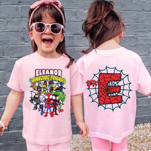 May include: A light pink short-sleeve t-shirt featuring a graphic print of cartoon characters and the text "Eleanor and Amazing Friends" on the front, and a large red letter "E" inside a black spiderweb design on the back of the garment.