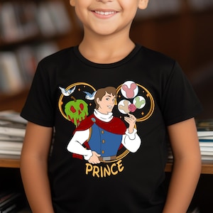 May include: Black t-shirt featuring a graphic of a prince holding a rose and an apple. The graphic is surrounded by a gold Mickey Mouse ear shape. The text "PRINCE" is below the graphic.