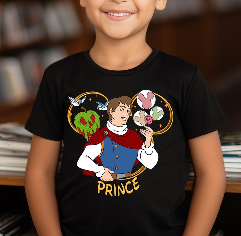May include: Black t-shirt featuring a graphic of a prince holding a rose and an apple. The graphic is surrounded by a gold Mickey Mouse ear shape. The text "PRINCE" is below the graphic.