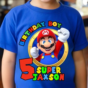 May include: A bright blue short-sleeved t-shirt featuring a graphic of Mario inside a gold ring. The text reads Birthday Boy, the number 5, and the name Super Jaxson in colorful block letters. The design includes a red hat and blue overalls.