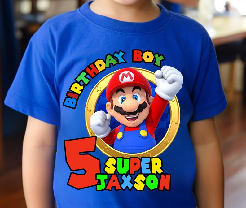May include: A bright blue short-sleeved t-shirt featuring a graphic of Mario inside a gold ring. The text reads Birthday Boy, the number 5, and the name Super Jaxson in colorful block letters. The design includes a red hat and blue overalls.