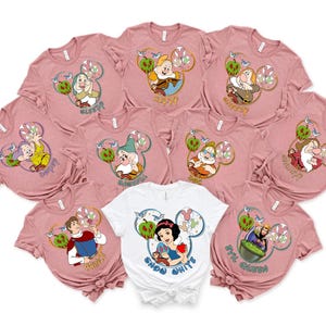 May include: Set of ten pink t-shirts featuring different Disney characters from Snow White. Each shirt depicts a different character with a Mickey Mouse ear design and text. The text on the shirts includes "Sleepy", "Happy", "Grumpy", "Dopey", "Sneezy", "Bashful", "Doc", "Evil Queen", "Snow White", and "Prince".