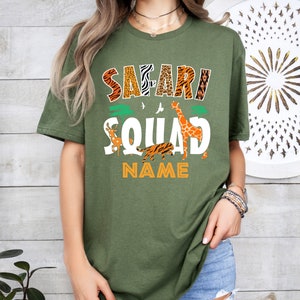 以下が含まれることがあります： オレンジ、茶色、黒の文字で「Safari Squad」と書かれた緑色のTシャツ。テキストはキリン、トラ、鹿に囲まれています。「NAME」というテキストは、動物の下にオレンジ色の文字で書かれています。