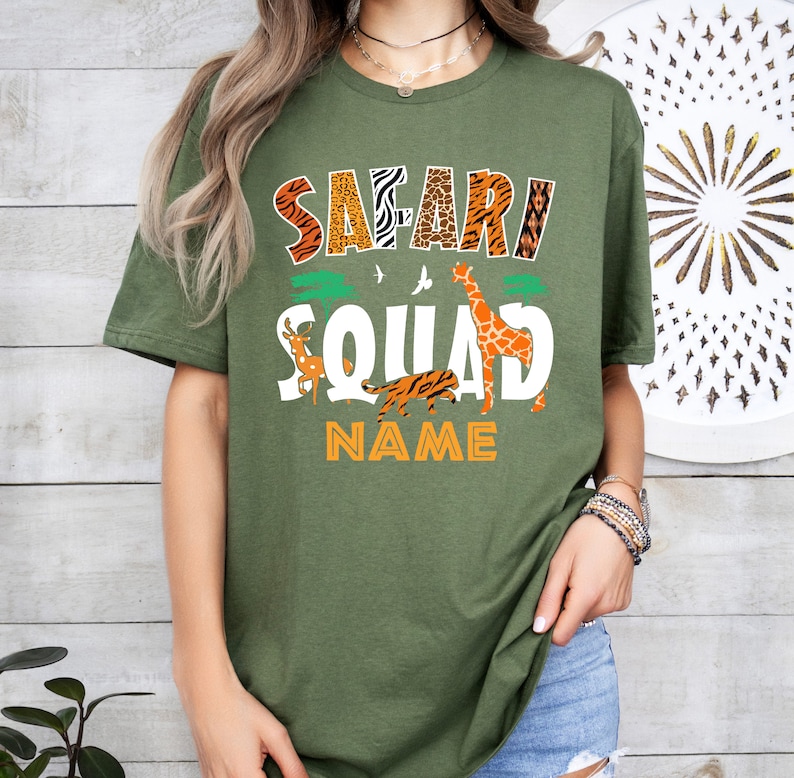 以下が含まれることがあります： オレンジ、茶色、黒の文字で「Safari Squad」と書かれた緑色のTシャツ。テキストはキリン、トラ、鹿に囲まれています。「NAME」というテキストは、動物の下にオレンジ色の文字で書かれています。