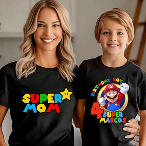 May include: Two black t-shirts with graphic prints. One shirt features the text "SUPER MOM" in red, yellow, and blue with a yellow star. The other shirt features a graphic of Mario, the text "BIRTHDAY BOY", the number "4", and "SUPER MARCOS".
