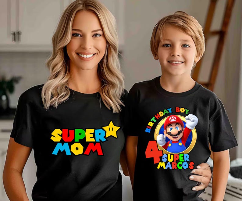 May include: Two black t-shirts with graphic prints. One shirt features the text "SUPER MOM" in red, yellow, and blue with a yellow star. The other shirt features a graphic of Mario, the text "BIRTHDAY BOY", the number "4", and "SUPER MARCOS".