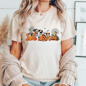 May include: A cream-colored short-sleeve t-shirt featuring a graphic print of cartoon characters dressed in Halloween costumes, sitting inside orange pumpkins. The design includes spider webs, bats, and various jack-o'-lanterns in a festive style.