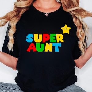 May include: A black t-shirt featuring the text SUPER AUNT in a multi-colored, blocky font style. The letters are colored in blue, red, green, and yellow. A yellow five-pointed star with two eyes is positioned to the right of the text.