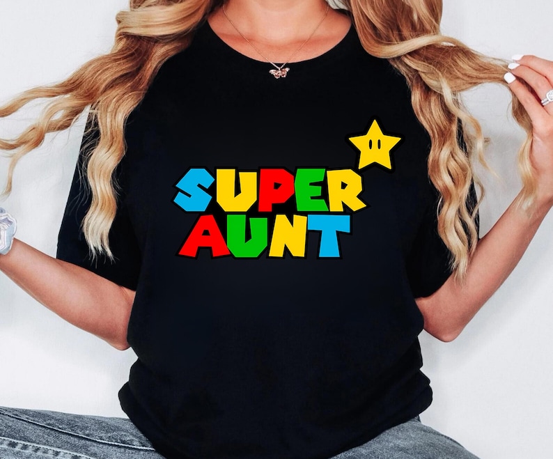 May include: A black t-shirt featuring the text SUPER AUNT in a multi-colored, blocky font style. The letters are colored in blue, red, green, and yellow. A yellow five-pointed star with two eyes is positioned to the right of the text.