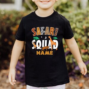以下が含まれることがあります： 「サファリ・スクワッド」と白文字で書かれた黒いTシャツ。キリン、シマウマ、ライオン、オリックスが描かれている。小さな木と鳥も描かれている。文字の下には「NAME」と書いてある。
