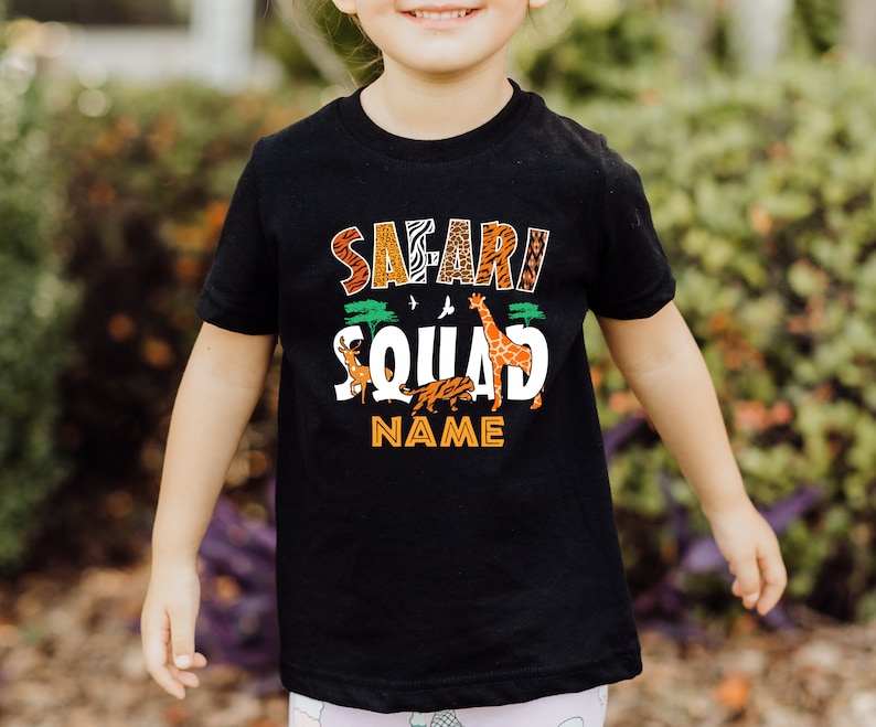 以下が含まれることがあります： 「サファリ・スクワッド」と白文字で書かれた黒いTシャツ。キリン、シマウマ、ライオン、オリックスが描かれている。小さな木と鳥も描かれている。文字の下には「NAME」と書いてある。