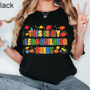 May include: A black short-sleeved t-shirt featuring the colorful text THIS IS MY LEGO BUILDING SHIRT in a blocky font. The design is accented with small, multi-colored plastic building brick graphics scattered around the lettering.