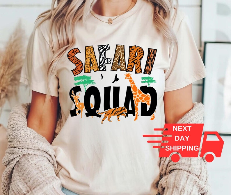 以下が含まれることがあります： 白いTシャツに、黒いグラフィックで「サファリ・スクワッド」と書かれており、キリン、シマウマ、ライオン、シカがジャングルの風景の中に描かれています。