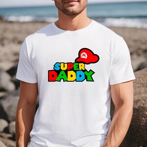 May include: A white short-sleeved t-shirt featuring a graphic print on the chest. The design displays the text "SUPER DADDY" in a multi-colored, blocky font with a red cap featuring a white "M" symbol positioned above the word "SUPER".