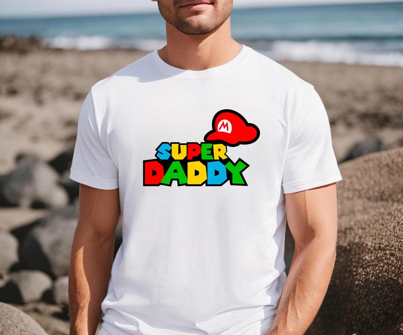 May include: A white short-sleeved t-shirt featuring a graphic print on the chest. The design displays the text "SUPER DADDY" in a multi-colored, blocky font with a red cap featuring a white "M" symbol positioned above the word "SUPER".