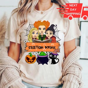 Puede incluir: Una camiseta de manga corta de color crema con un gr&aacute;fico de tres brujas de dibujos animados, una telara&ntilde;a, una calabaza, un caldero morado y un gato negro con sombrero de bruja. Una pancarta naranja muestra el texto CUSTOM NAME.