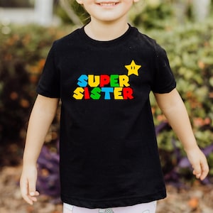May include: A black short-sleeved t-shirt featuring the text SUPER SISTER in a multi-colored block font with a yellow star graphic positioned above the word SISTER. The design is centered on the chest of the shirt.