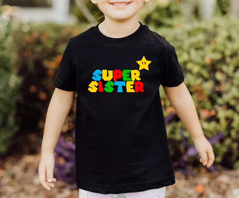 May include: A black short-sleeved t-shirt featuring the text SUPER SISTER in a multi-colored block font with a yellow star graphic positioned above the word SISTER. The design is centered on the chest of the shirt.
