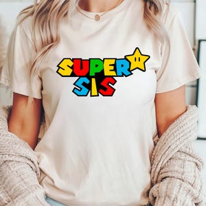May include: A cream-colored short-sleeve t-shirt featuring the text SUPER SIS in a bold, blocky, multi-colored font. The letters are colored in yellow, red, and blue, and a yellow five-pointed star icon is positioned to the right of the text.