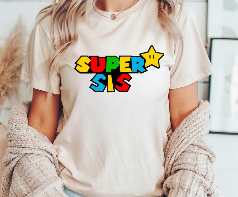 May include: A cream-colored short-sleeve t-shirt featuring the text SUPER SIS in a bold, blocky, multi-colored font. The letters are colored in yellow, red, and blue, and a yellow five-pointed star icon is positioned to the right of the text.