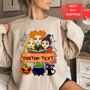 Puede incluir: Una sudadera beige de cuello redondo con un estampado gr&aacute;fico de tres brujas de dibujos animados con cabello naranja, rubio y negro, un fondo de telara&ntilde;a, una calabaza, un caldero morado y un gato negro con un sombrero a rayas y texto.