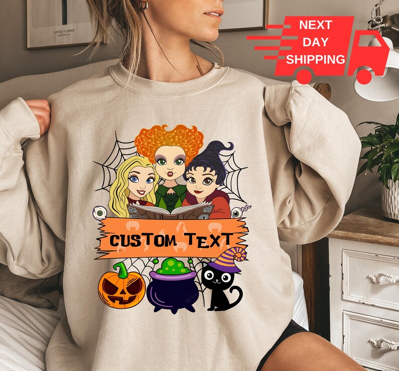 Puede incluir: Una sudadera beige de cuello redondo con un estampado gr&aacute;fico de tres brujas de dibujos animados con cabello naranja, rubio y negro, un fondo de telara&ntilde;a, una calabaza, un caldero morado y un gato negro con un sombrero a rayas y texto.