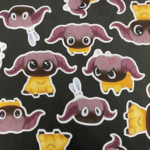 May include: A collection of cartoon stickers featuring a horned creature with large eyes. The stickers have a color palette of purple, brown, yellow, and white. Some stickers have a small bunny or star accents. The stickers are on a black background.