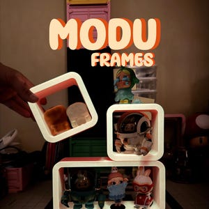May include: White and pink square display frames holding various figurines. The frames are arranged in a vertical stack, with the text "MODU FRAMES" in orange and white above. The figurines include a space-suited figure, a bunny, and other characters.