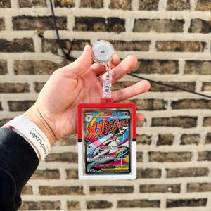 May include: A red and white badge holder with a retractable reel, holding a Pokémon trading card. The card features Japanese text and a colourful illustration of a creature. The badge holder is held against a brick wall.