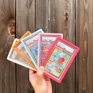 May include: A collection of five graded trading cards in colourful plastic frames, including white, orange, light blue, pink, and magenta. Each card is encased in a protective slab with a PSA label at the top, displaying various Pokémon illustrations.
